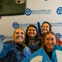 Group photo: someone taking a selfie in front of GVSU banner, the group posing with foam fingers and the GV signs